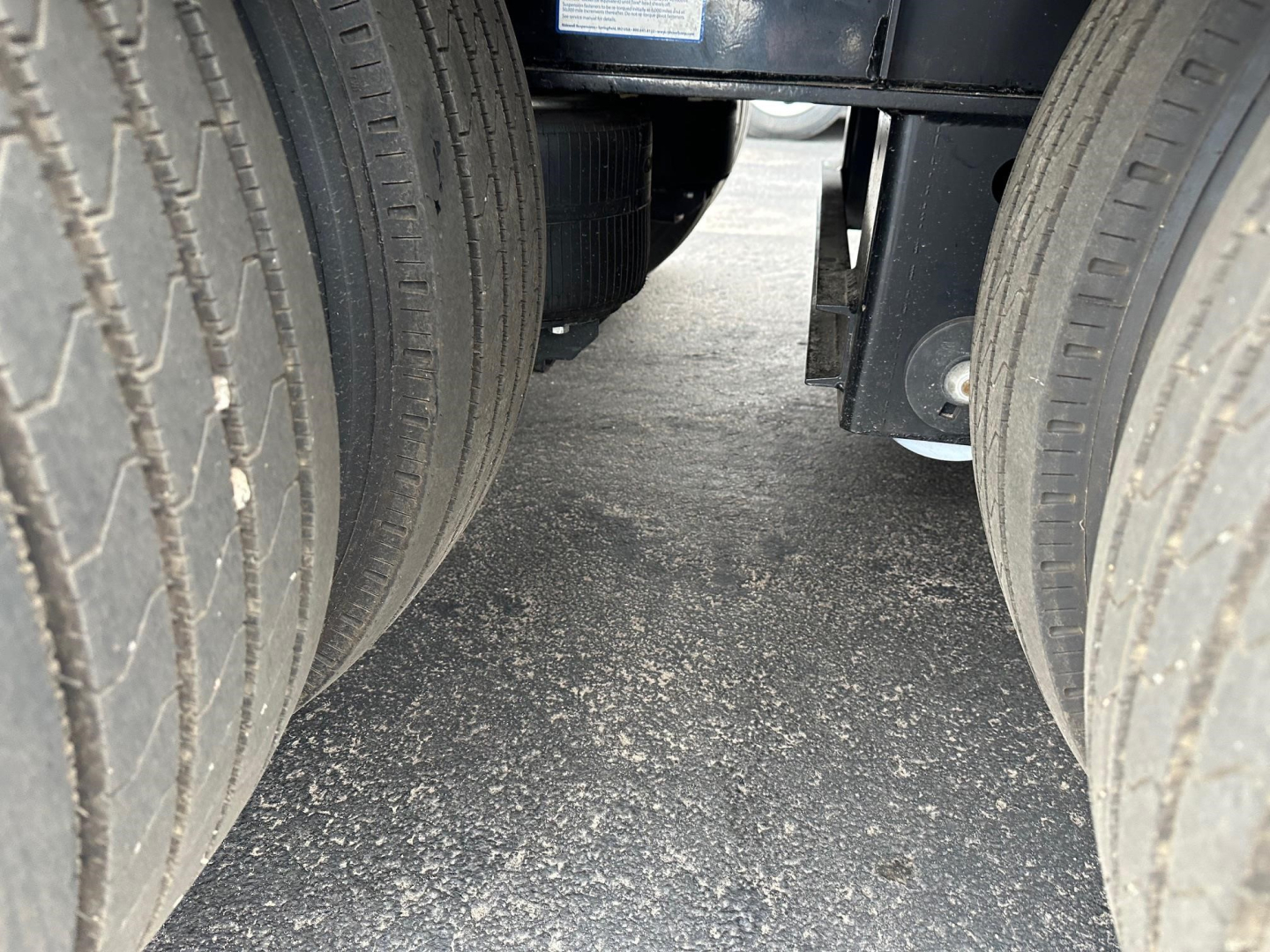 Trailer dual rear tires with undercarriage and part of frame visible between wheels; asphalt surface beneath.