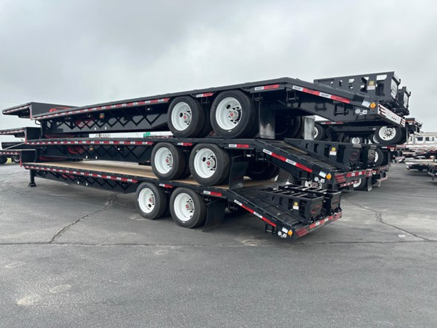 Trailer stack showing three fold-out steel drop decks with dual rear axle wheels and visible tires, black frame and reflective safety tape.