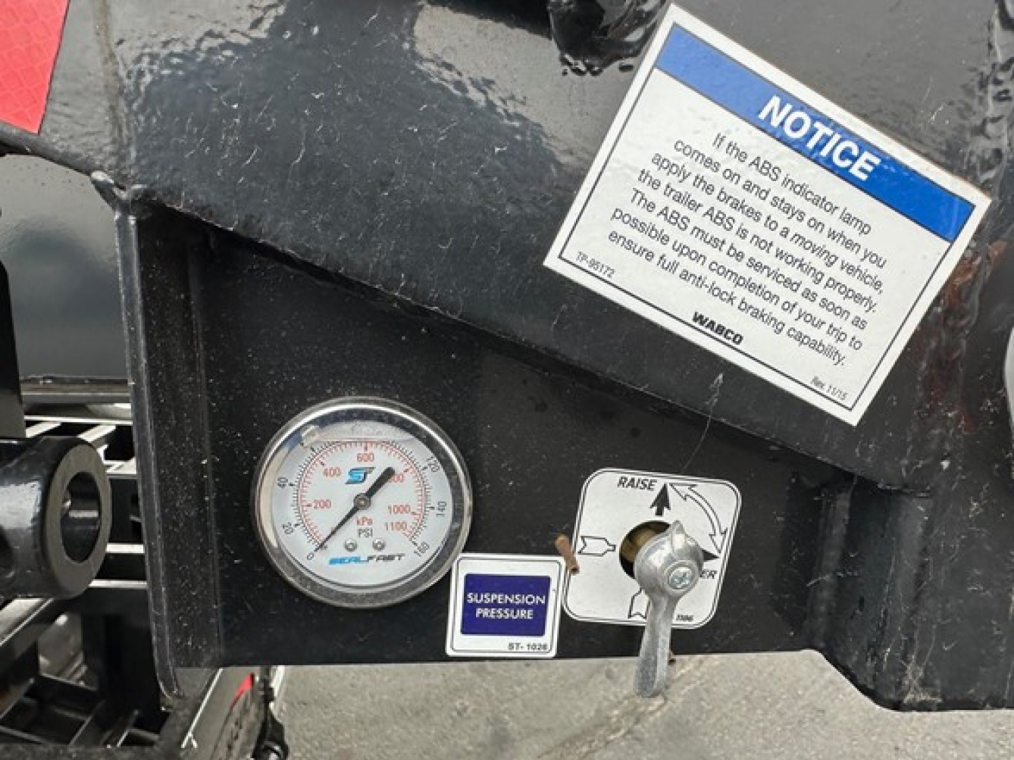 Drop deck trailer front panel with round pressure gauge and suspension control knob. Notice label with instructions and a blue notice card on the black metal surface.