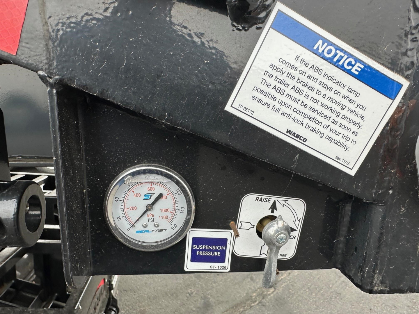 Trailer dashboard close-up showing gauges and label: suspension pressure gauge, blue warning label, and notice sticker on black metal surface.