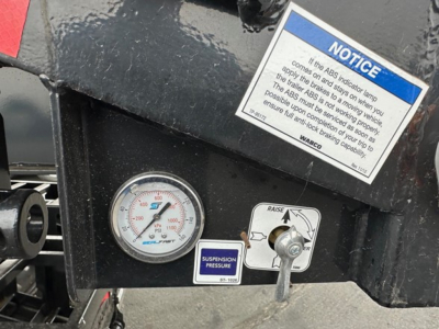 Dashboard area of a trailer showing a round pressure gauge, a blue notice sticker, a suspension pressure gauge label, and a control lever with a white label reading 'RAISE' and arrows.