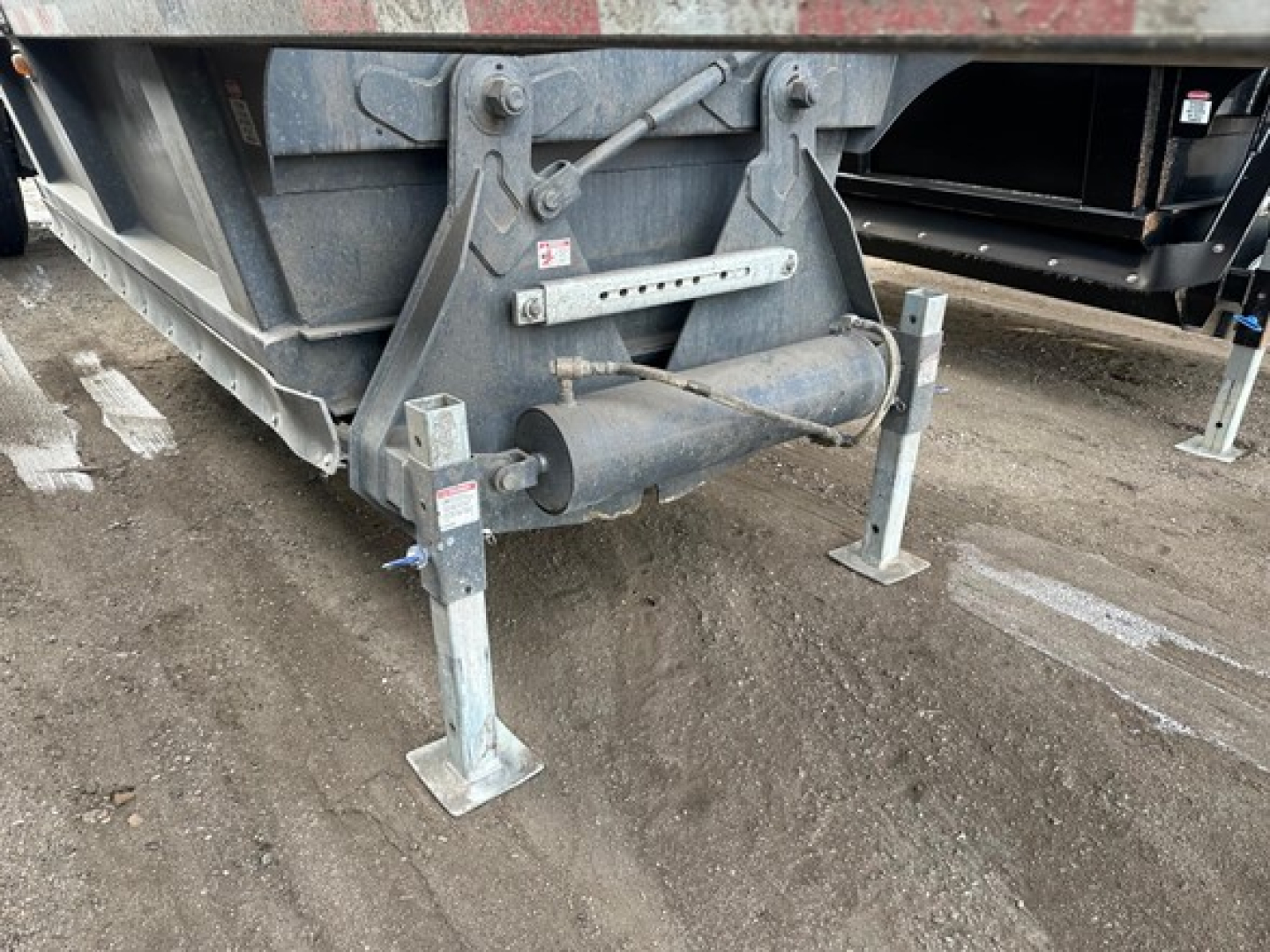 Trailer undercarriage showing landing gear legs and a drum-style PTO or hub with hydraulic components and warning stickers
