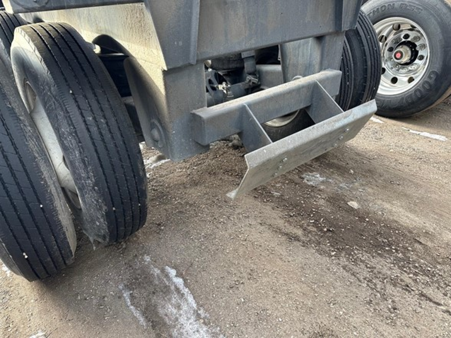 Rear view of a bottom-dump trailer with dual rear axles, showing the undercarriage and hydraulic dumping mechanism. Tires and parking area visible.