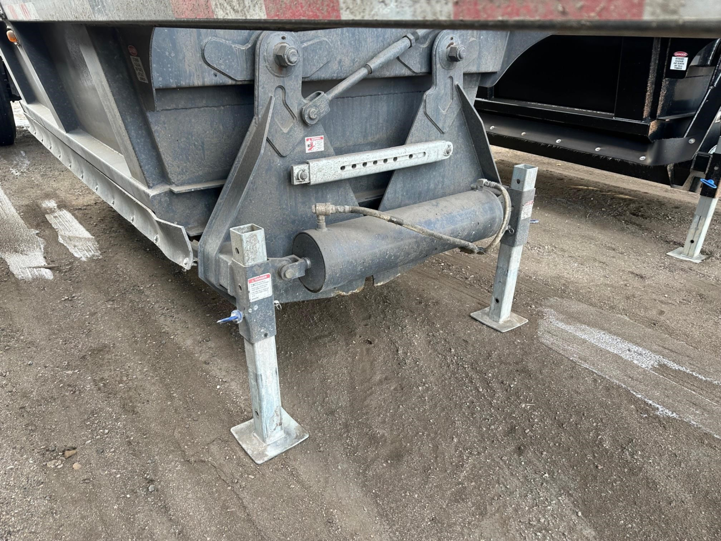 Bottom dump trailer tongue area showing support legs and hydraulic cylinder assembly with warning labels and frame crossmember.