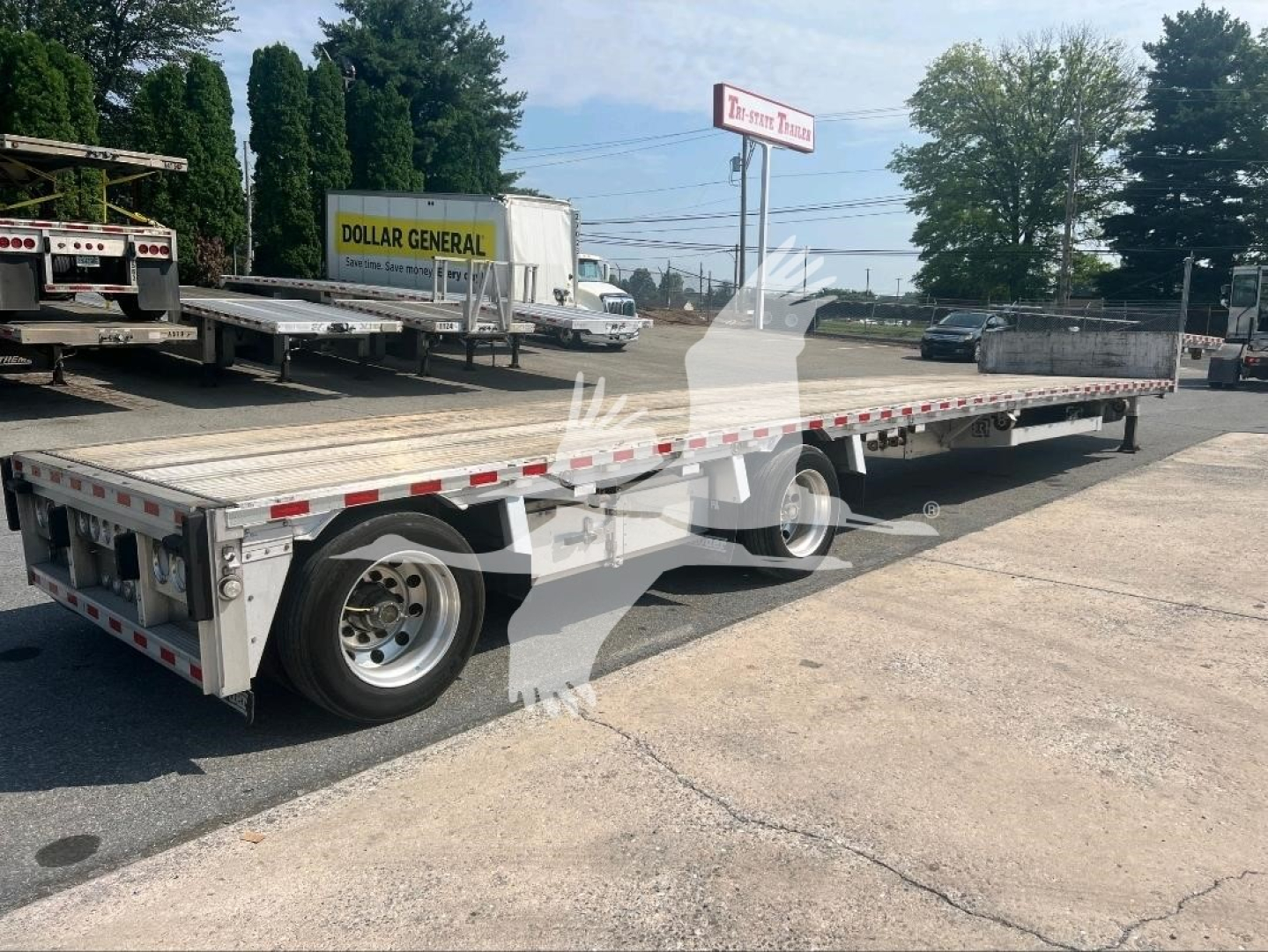 Flatbed trailer in a lot with other trailers; silver aluminum deck, long dropdeck with dual axles and wheels visible; Dollar General sign in background.