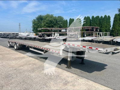 Dropdeck trailer on an empty lot, with another trailer in background, showing dual-axle platform and support legs.