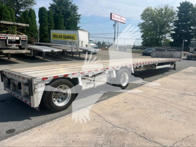 Flatbed trailer in a dealership lot showing long drop-deck trailer with dual rear axles and red/white reflective tape along sides.