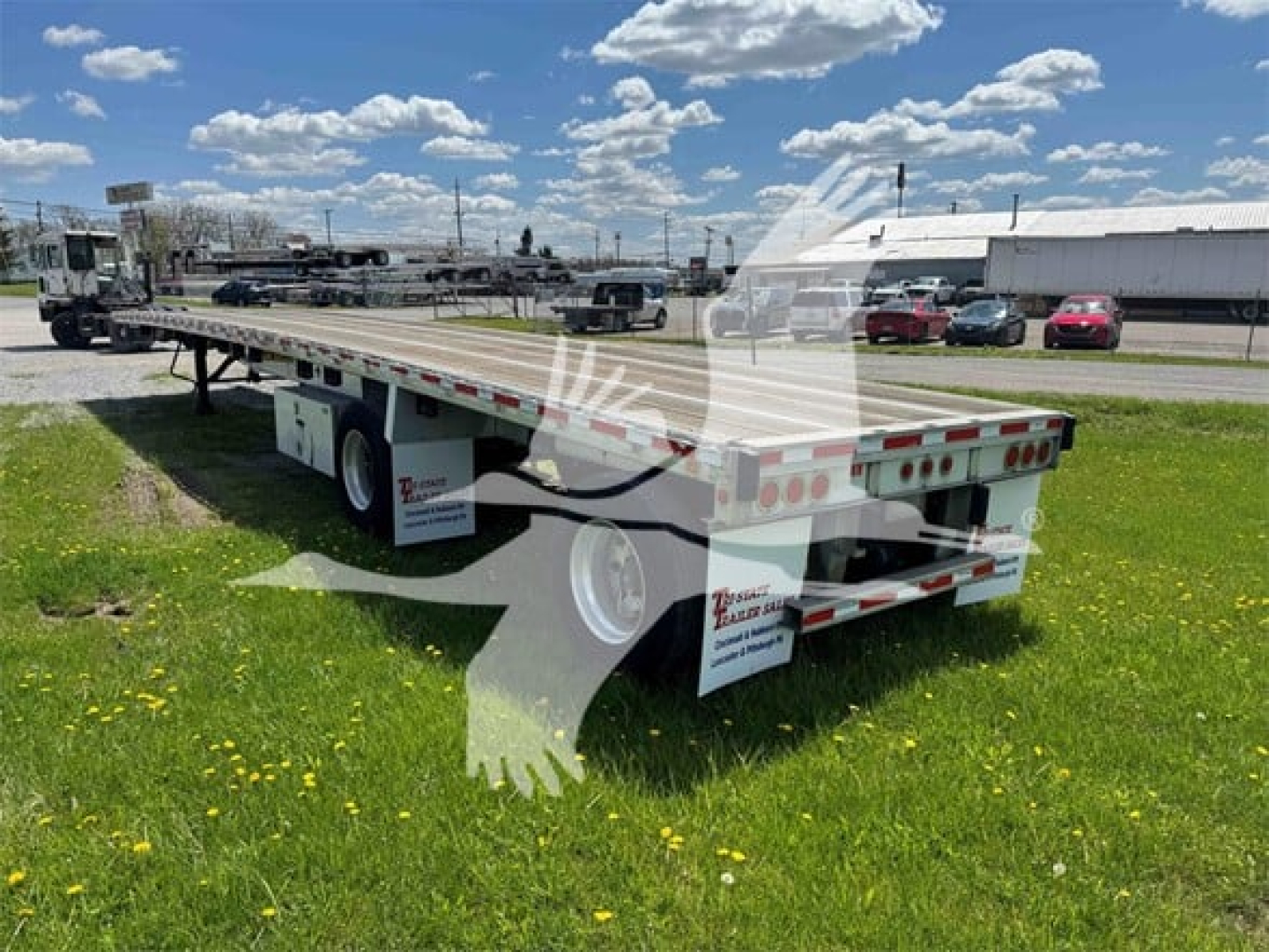 Flatbed trailer showing rear and left side with dual rear axles, reflective tape, and mudflaps; signage on side panels; blue sky with clouds.
