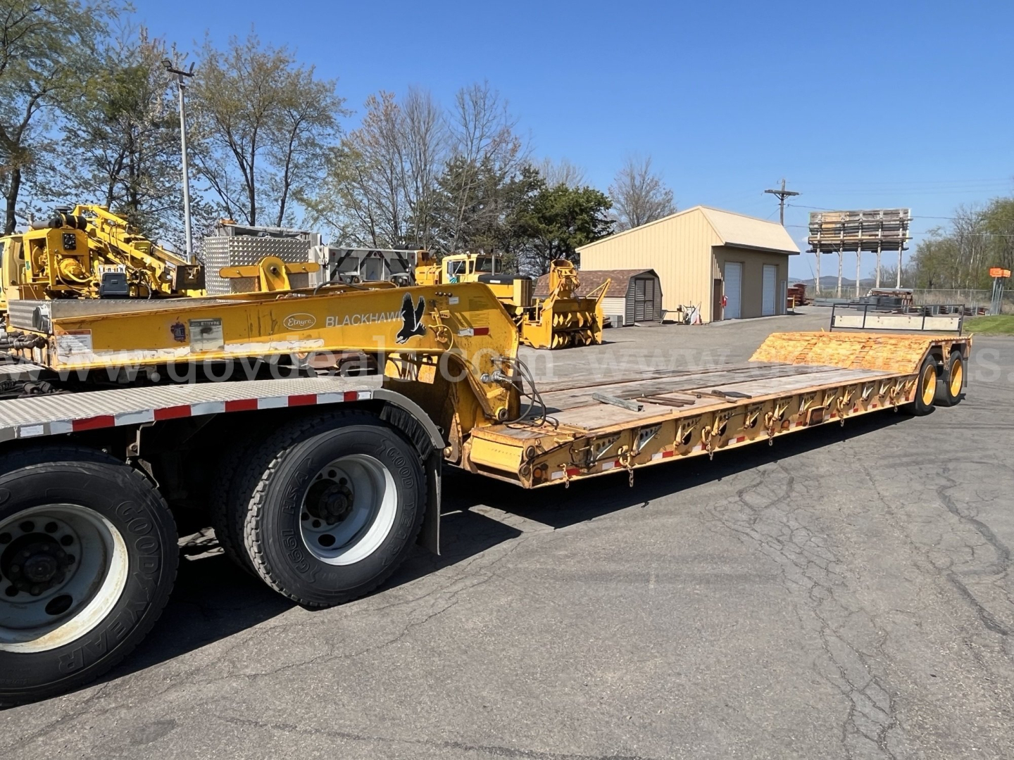 Lowboy trailer with yellow equipment on a flatbed; side shows 'LOAD' branding and black stripes; clear view of long deck and dual rear axles; surrounding buildings and trees.