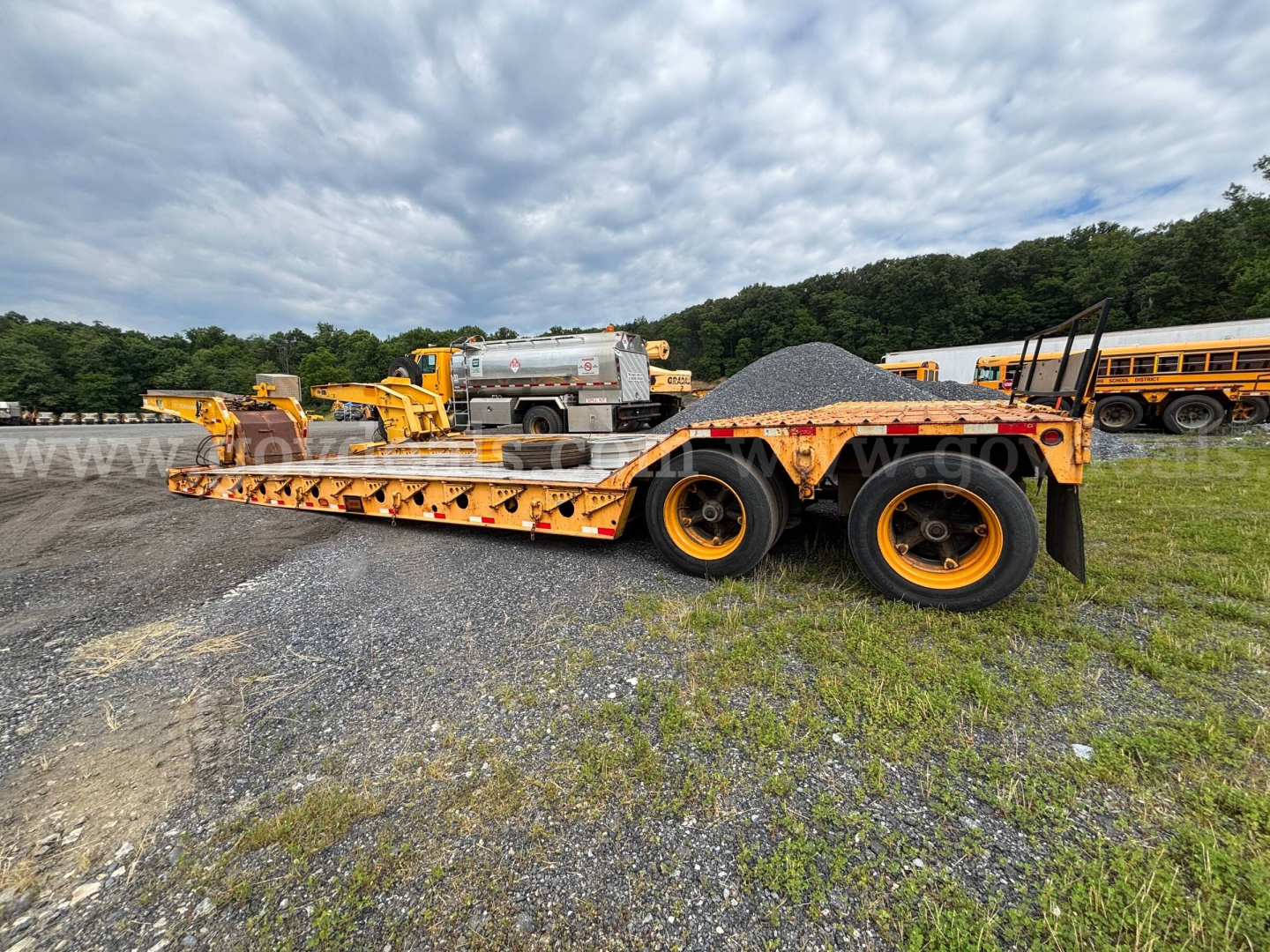 Lowboy trailer with long flatbed and multiple axles, parked on gravel; distant orange equipment and a silver tanker behind.