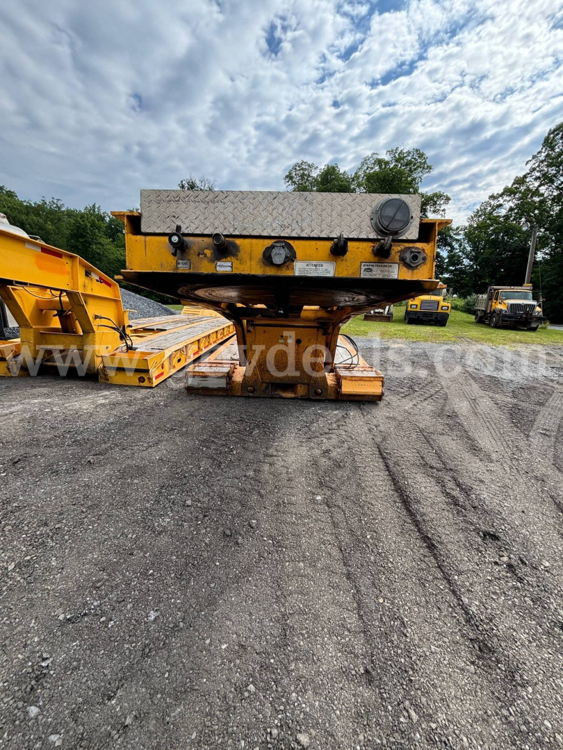 Lowboy trailer in service showing metal control panel with knobs and gauge and diamond plate box on top, yellow equipment on gravel lot.