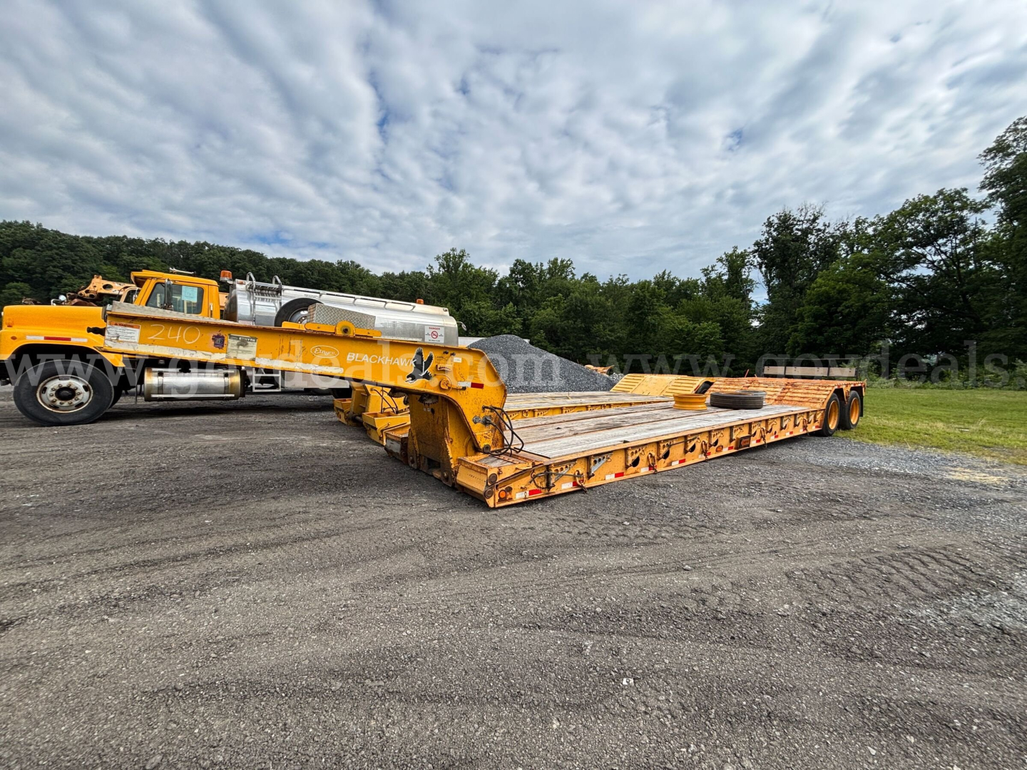 Lowboy trailer with yellow frame and long flatbed, front gooseneck and multiple axles, parked on a gravel lot with a truck in the background.