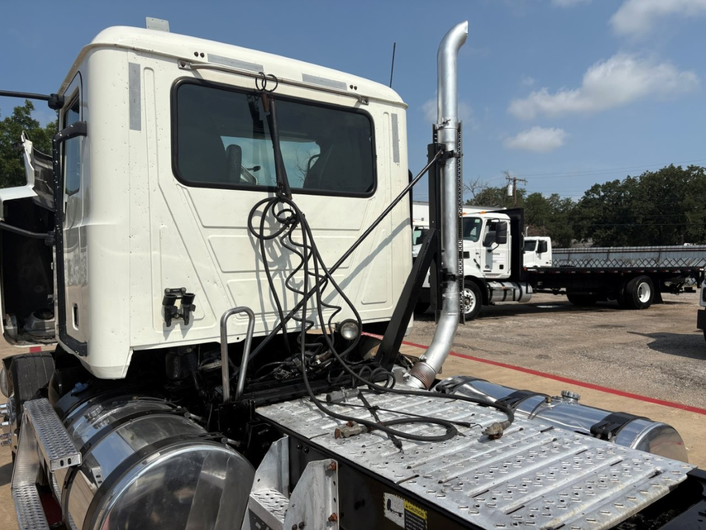 All-white Mack PI64T cab with chrome exhaust stack and truck cab, exposed battery cables, and aluminum fuel tanks. View of front-left side and hood, with other trucks in background.
