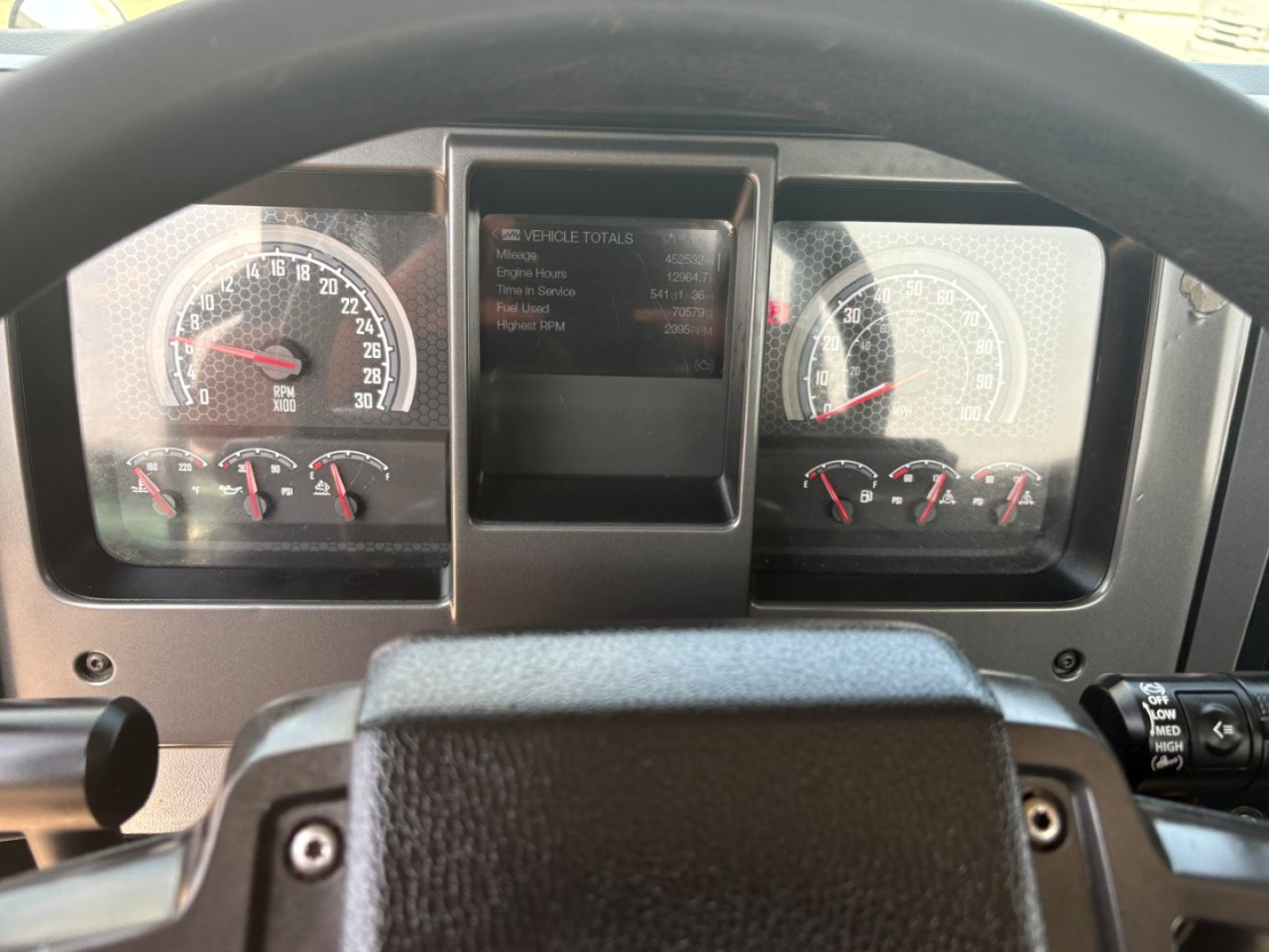 Dashboard of a Mack PI64T truck showing instrument cluster and center display. Gauges include speedometer, tachometer, fuel and coolant gauges. Center screen appears to list vehicle totals and engine hours. Steering wheel in foreground.