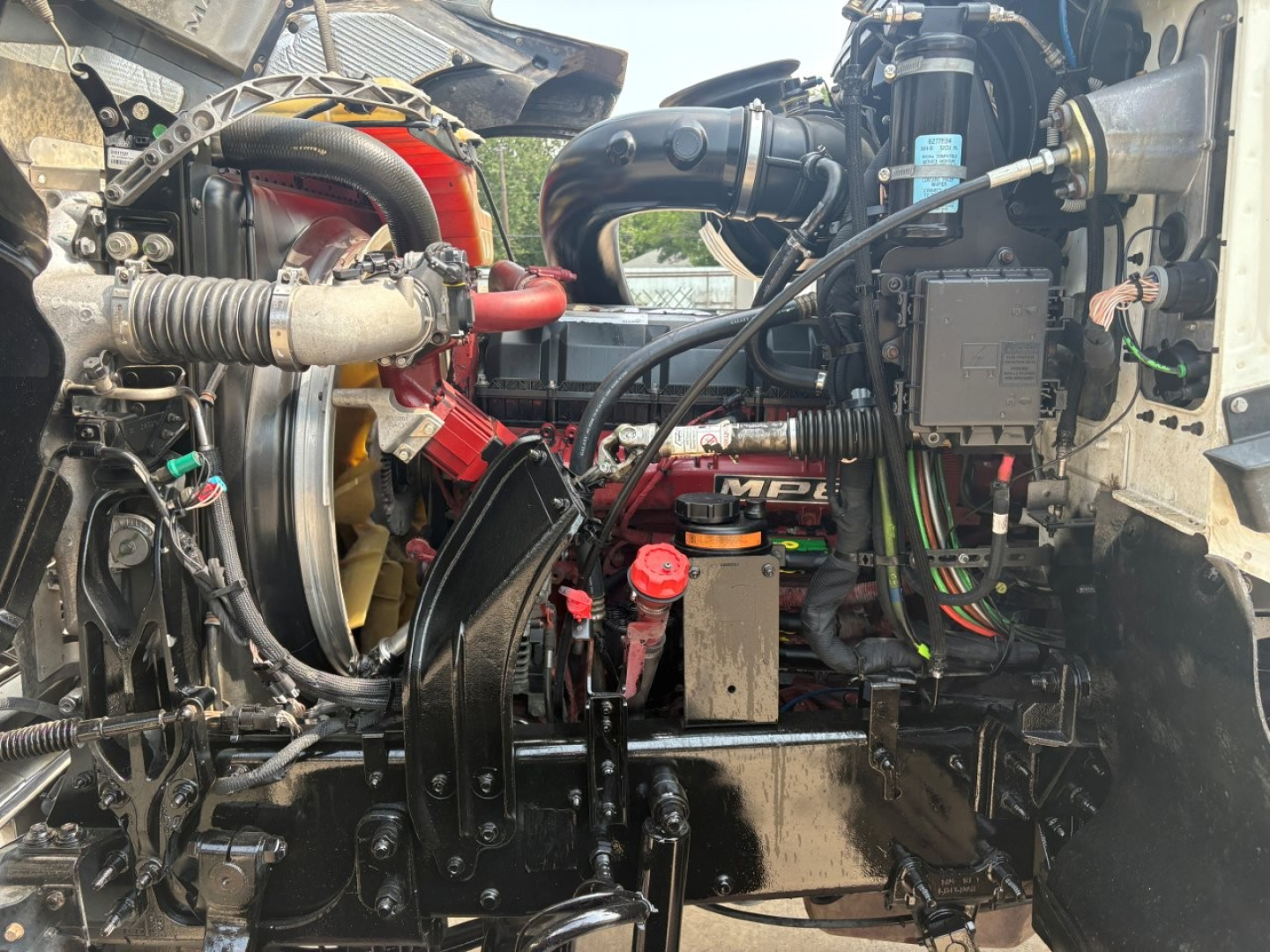 Engine bay of a 2020 Mack PI64T conventional daycab showing engine components, hoses, coolant reservoir, wiring, air intake, and radiator area.