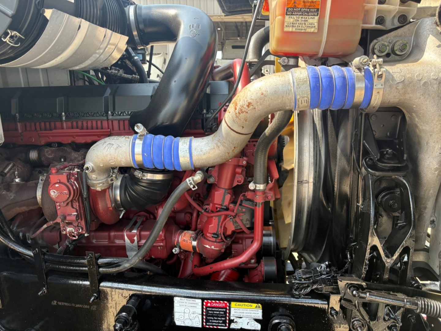 Mack PI64T engine bay showing red engine components, blue silicone hose clamps, radiator, fan and coolant reservoir with warning label.