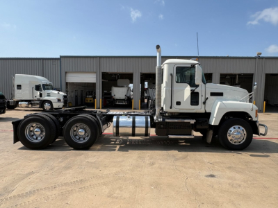 Truck side view of a 2020 Mack PI64T conventional daycab showing dual rear axles, chrome fuel tank, exhaust stack, and white cab against a warehouse background.