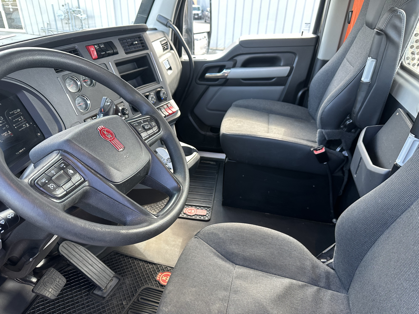 Kenworth T680 cab interior showing steering wheel and driver seat with dashboard gauges.