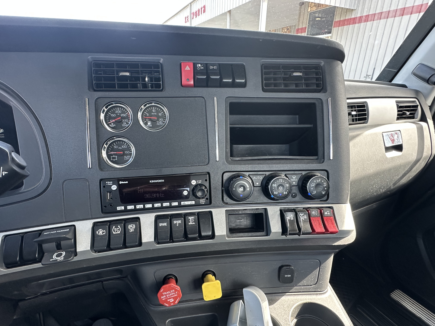 Dashboard view of a 2022 Kenworth T680 conventional daycab showing gauge cluster, radio, climate controls, hazard switch, and various switches.