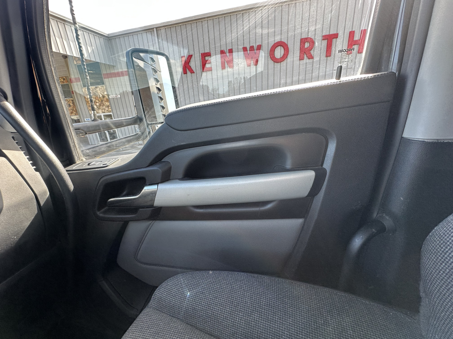 Kenworth T680 interior door panel view from the passenger seat showing window, armrest, and handle with Kenworth dealership signage reflected in the window.