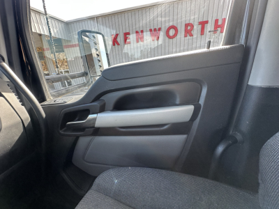 Kenworth T680 interior door panel view from the passenger seat showing window, armrest, and handle with Kenworth dealership signage reflected in the window.