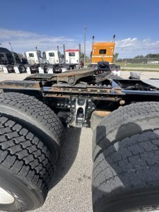 Dual rear wheel assembly of a semi truck chassis with fifth-wheel hitch and air suspension visible; background shows multiple truck cabs and an orange cab.