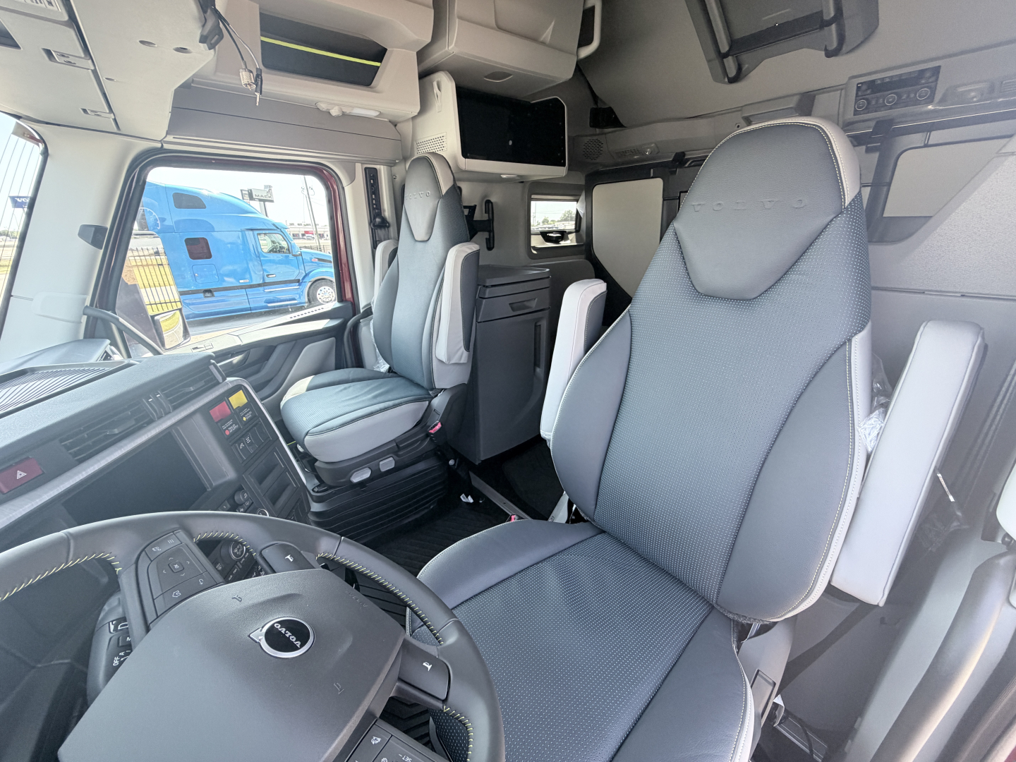 Cab interior of a Volvo VNL truck showing steering wheel, driver seat, passenger seat, overhead storage, and dashboard details.