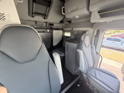 Cab interior of a Volvo truck with driver's seat and captain seats, overhead compartments, and a small monitor; visible text includes 'VOLVO' embossed on seat headrest.