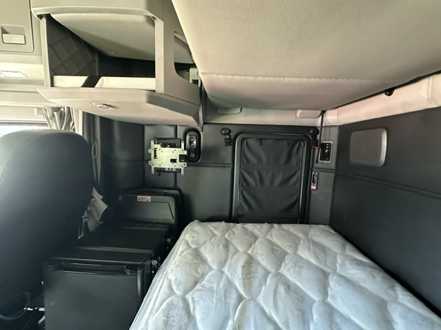 Kenworth T680 sleeper cab interior showing upper bunk area, storage shelf, and ladder/storage compartments with a quilted mattress in the back. Cab panel details and wiring visible.