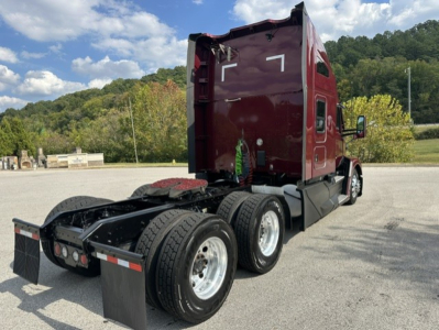 Kenworth T680 vermilion cab with sleeper, rear view showing dual rear axles and fifth wheel, flat road, trees in background.
