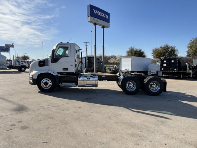 2027 Mack AN64T truck chassis in a dealership lot with a Volvo sign in background; side view shows cab, fuel tanks, dual rear axles, no trailer.