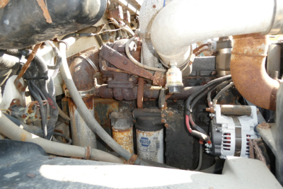 Engine bay of a used 1999 Freightliner FL70 showing hoses, rusted components, alternator, and fuel filters.
