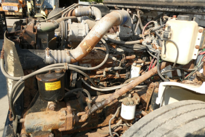 Engine bay of a Freightliner FL70 with exposed hoses, filters, and coolant reservoir; visible rusty metal components and a large intake pipe.