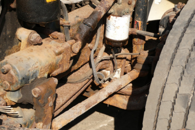 Diesel truck chassis showing rusted front axle and suspension components of a Freightliner FL70 with exposed hoses and a white filter canister.