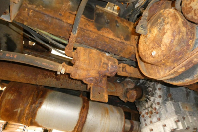 Rusty mechanical drivetrain components of a Freightliner FL70 undercarriage showing hydraulic lines, steering linkage, and a rusted transfer/axle assembly.