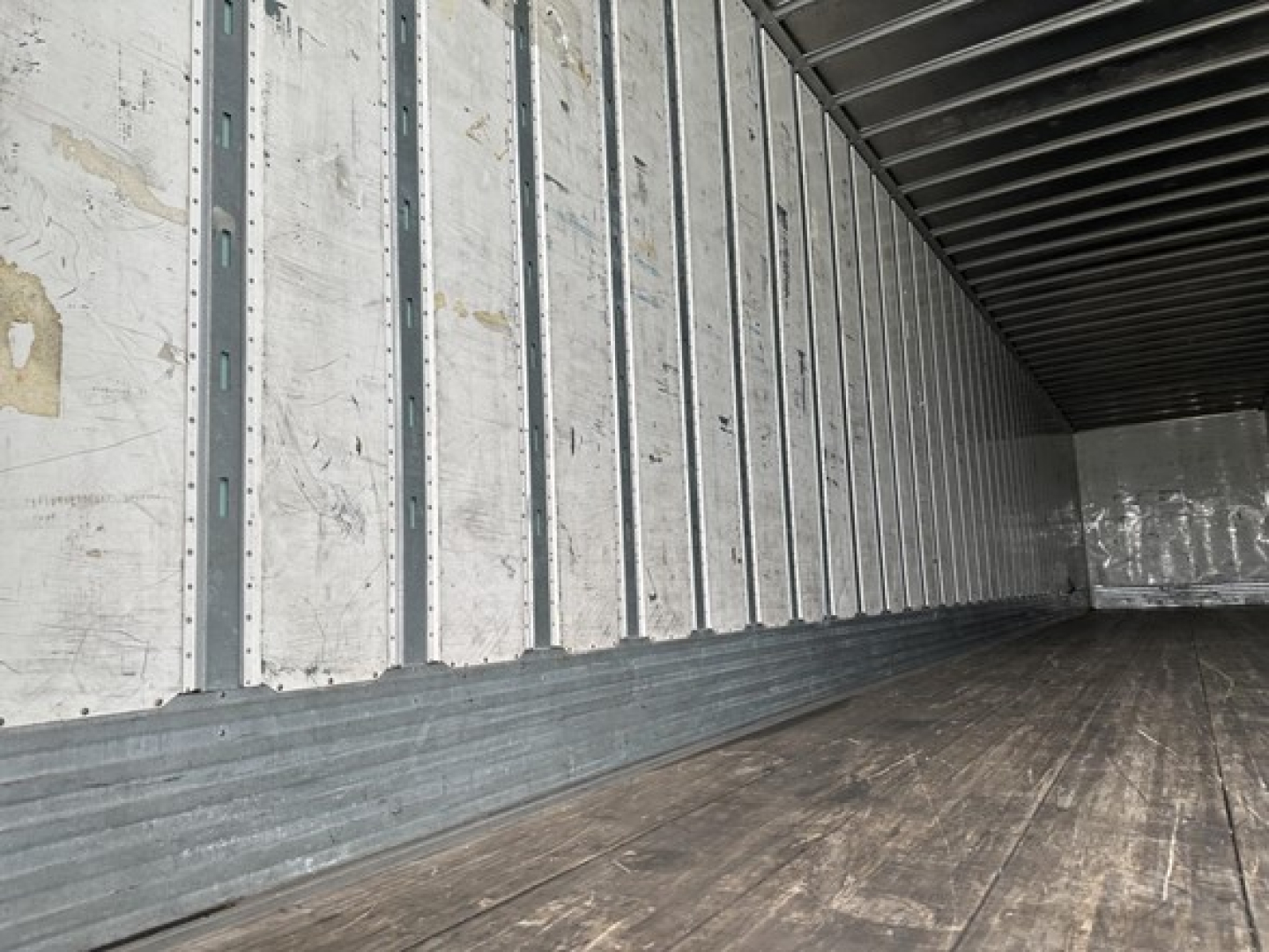 Trailer interior showing wooden floor and metal side walls with vertical studs, empty cargo area, light from open rear. No identifying text visible.