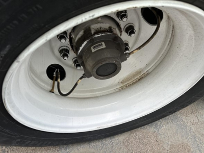 Wheel hub with white trailer rim, black hub cap, and tire showing mounting bolts and a label, viewed from side angle.