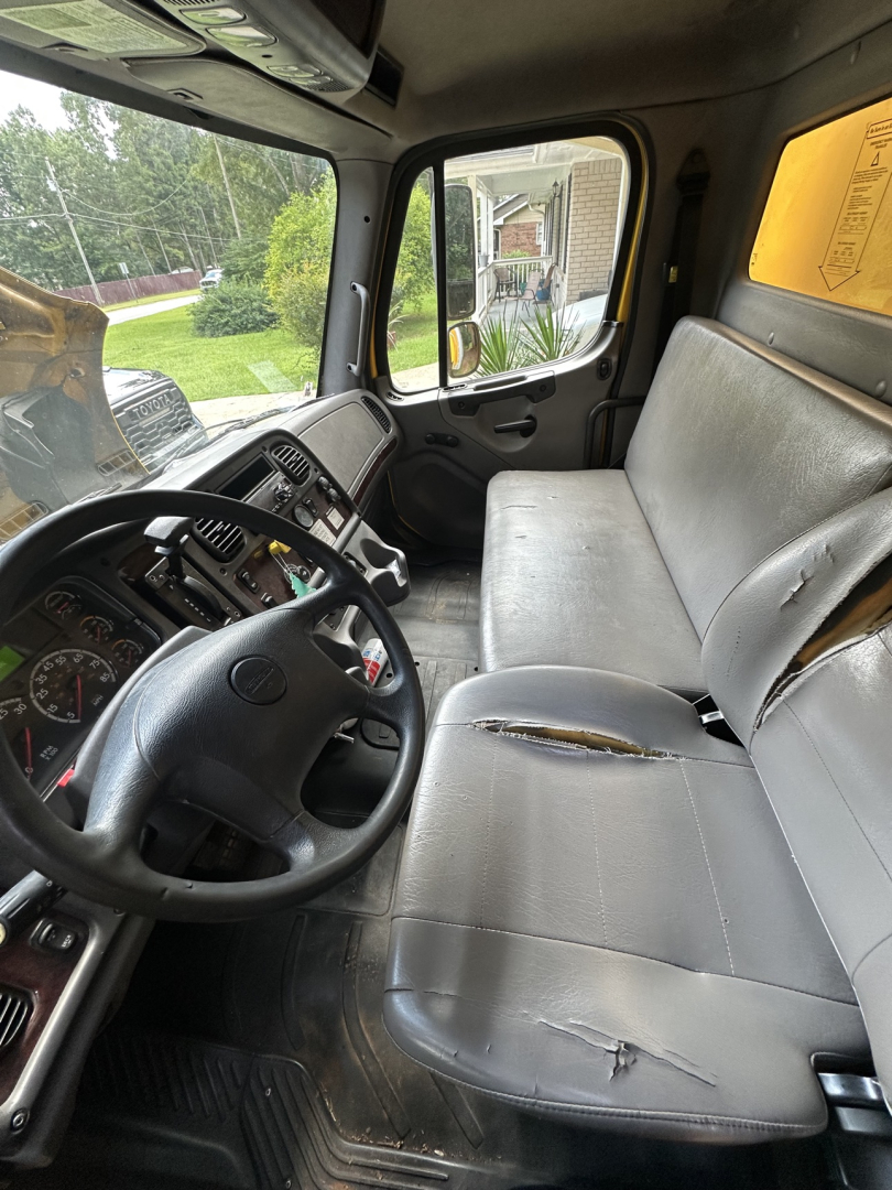 Cab interior of a 2019 Freightliner M2 106 showing the steering wheel, dashboard, and a worn grey bench seat with visible tears.