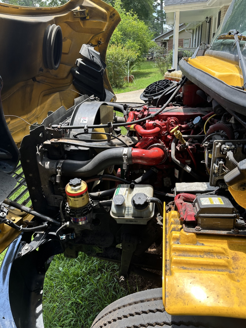 Truck engine bay of a 2019 Freightliner M2 106 showing red ignition hoses, yellow coolant reservoir, radiator with fan, battery and various fluid reservoirs.