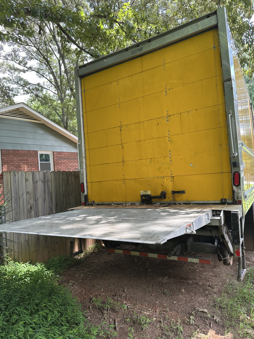Truck bed of a Freightliner M2 106 with a yellow drop-side cargo box, rear loading ramp lowered, visible VIN plate not shown, outdoor setting.
