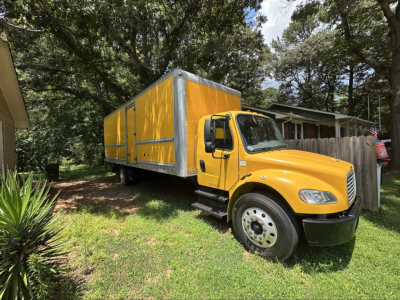 Yellow Freightliner M2 106 box truck parked on grass with green trees in background, large cargo box with yellow cover and silver frame, front cab visible.