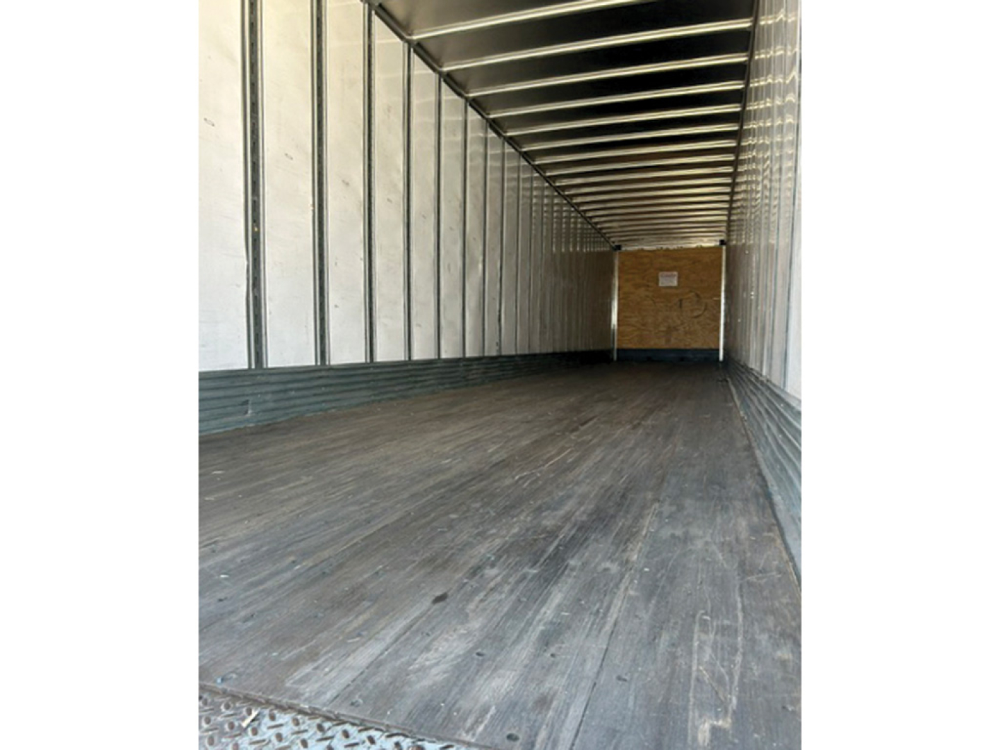 Trailer interior showing empty flatbed area with metal sidewalls and ceiling, wooden rear wall, and dark non-slip floor.