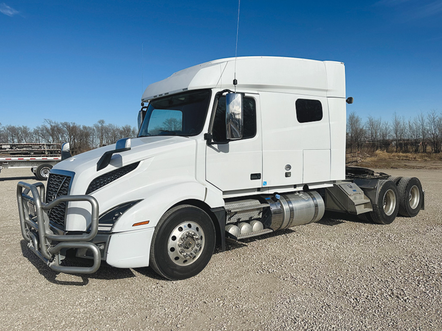 White Volvo VNL 740 semi-truck tractor with a high sleeper cabin, front grille guard, dual exhaust and side fuel tanks parked on gravel.