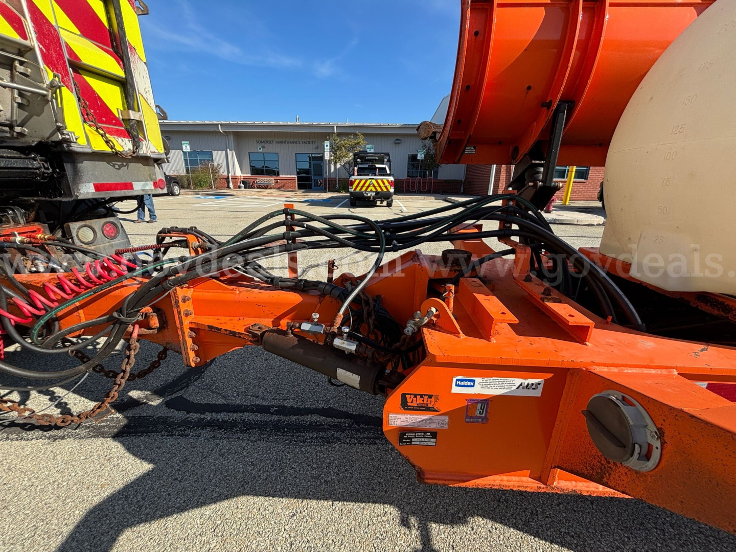 Front left view of a 2013 Mack GU Granite plow truck chassis with orange equipment and hydraulic hoses; VIN tag and stock number not visible; foreground showing hydraulic winch area and control components.