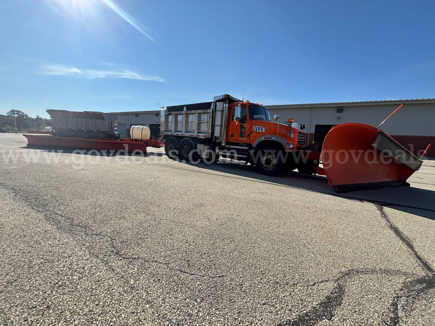 Orange Mack GU713 Granite highway plow truck with large snowplow blade attached in front, utility tank and equipment in bed, daytime.