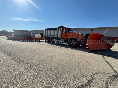 Orange Mack GU713 Granite highway plow truck with large snowplow blade attached in front, utility tank and equipment in bed, daytime.