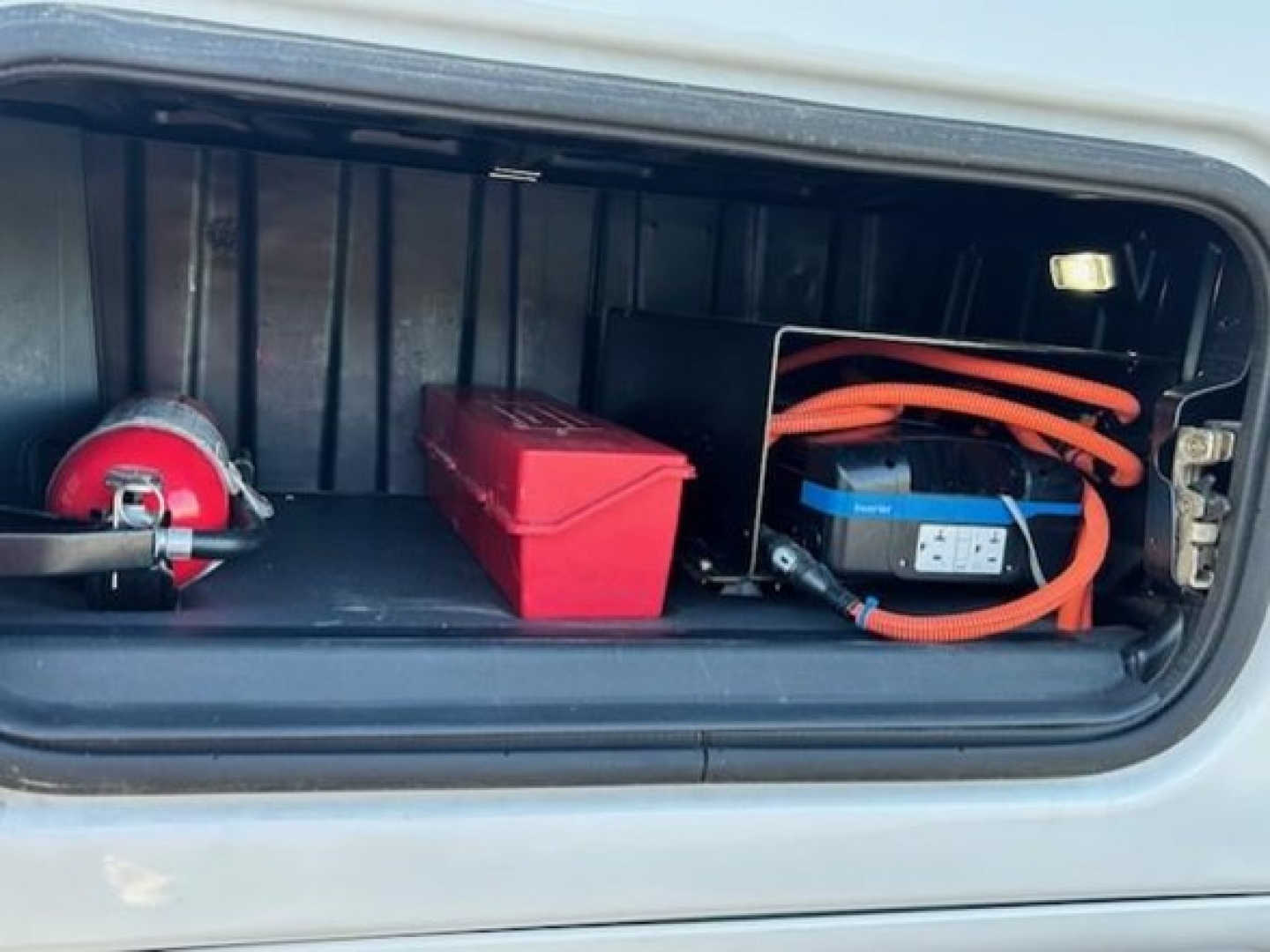 Truck front battery and storage compartment of a Freightliner Cascadia 126 sleeper showing blue AGM battery with orange cables, red toolbox, and red storage box.