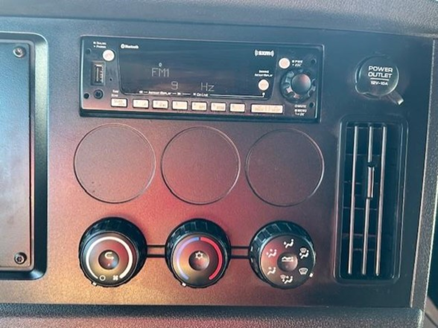Dashboard area of a Freightliner Cascadia showing radio/FM unit, three circular climate controls, and a power outlet knob with ventilation slot.