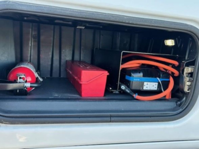 Truck front battery and storage compartment of a Freightliner Cascadia 126 sleeper showing blue AGM battery with orange cables, red toolbox, and red storage box.