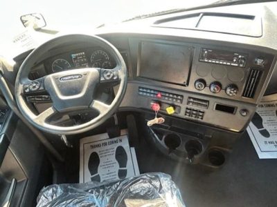 Cab interior of a Freightliner Cascadia shows steering wheel, dash with gauges, large central touchscreen, radio, climate controls, and ignition area.